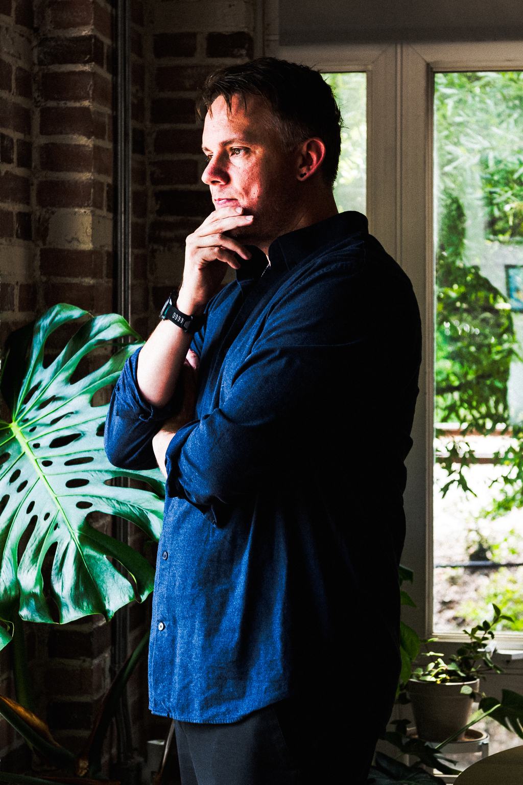 Christopher Shank standing at an open window beside a monstera, one hand at his chin.