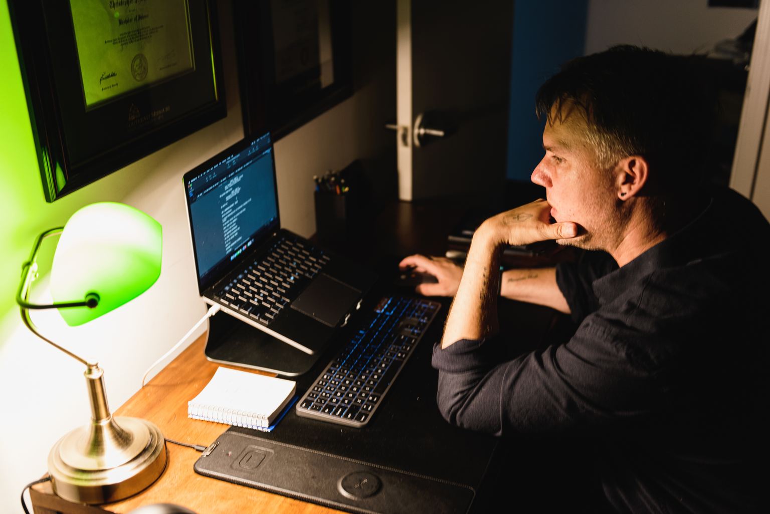 Christopher Shank seen over his shoulder at a night desk, laptop and notebook lit by a green banker’s lamp.