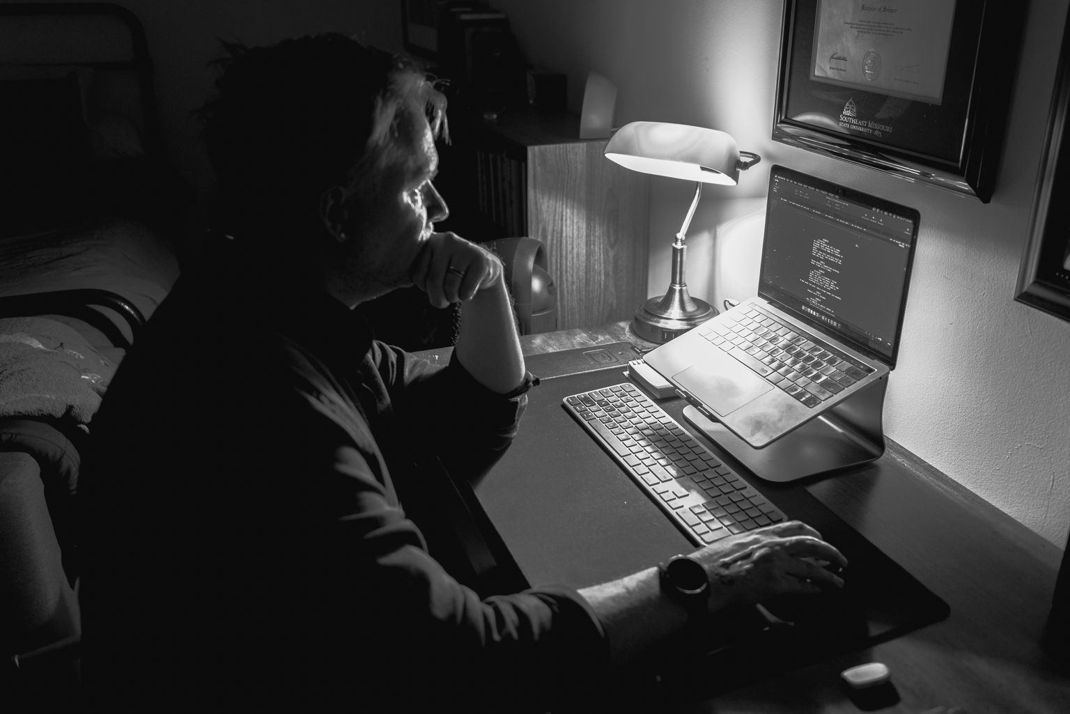 Christopher Shank at his desk in a dark room, one hand at his chin, reading his laptop screen.