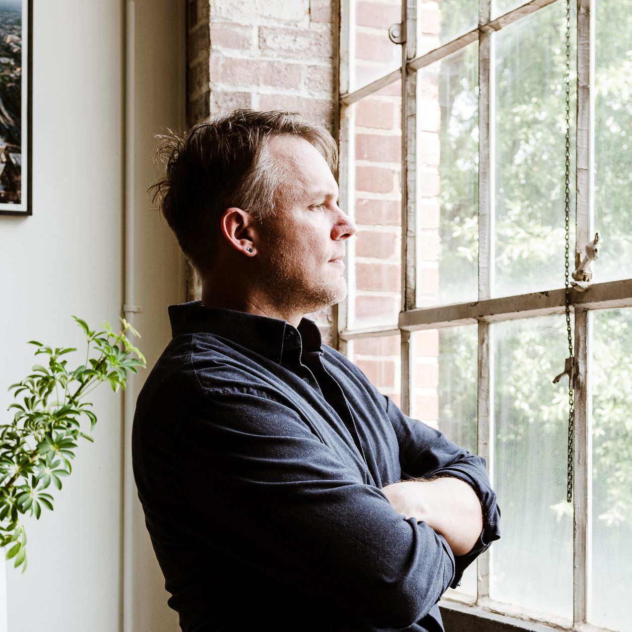 Portrait of Christopher Shank beside a tall factory window, arms crossed.